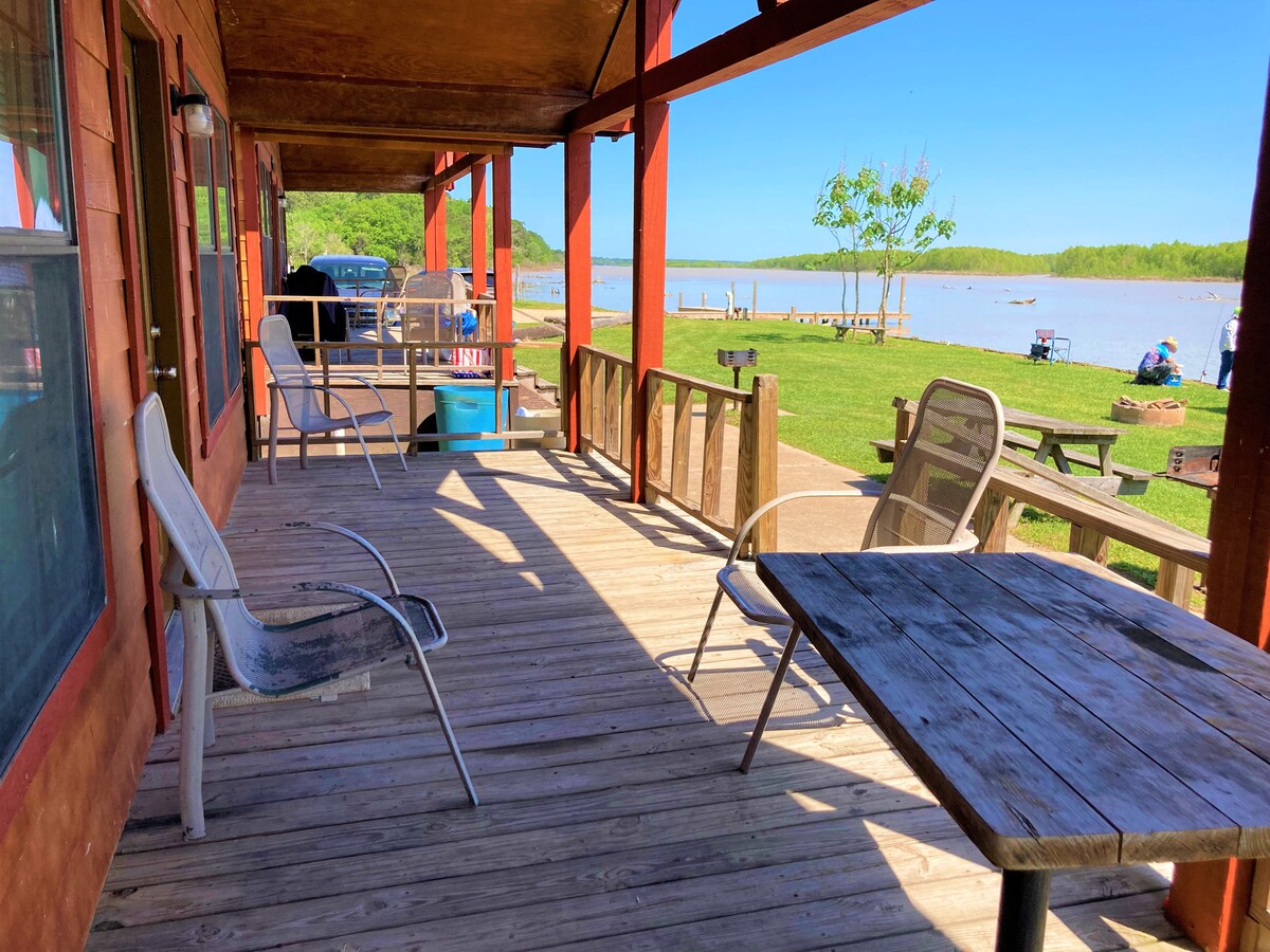 The spacious front porch features a wooden floor with a large picnic table and several seating options. A barbecue grill is positioned nearby, while a scenic view of the lake and surrounding greenery is visible in the background.