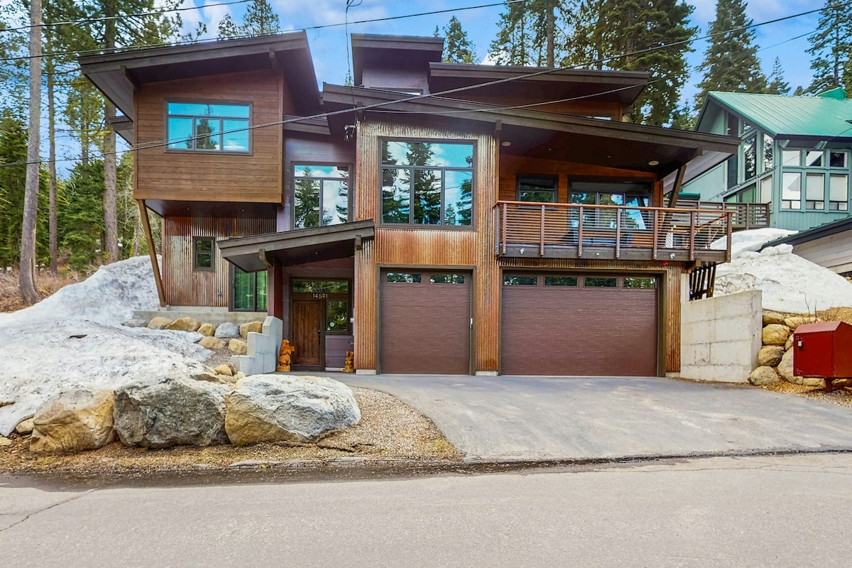 The exterior of Red Mountain Lodge features a modern design with natural wood siding and large windows. The home is positioned on a gently sloping lot, with snow patches visible around the property. Two garages are present at the front, complementing the clean driveway.