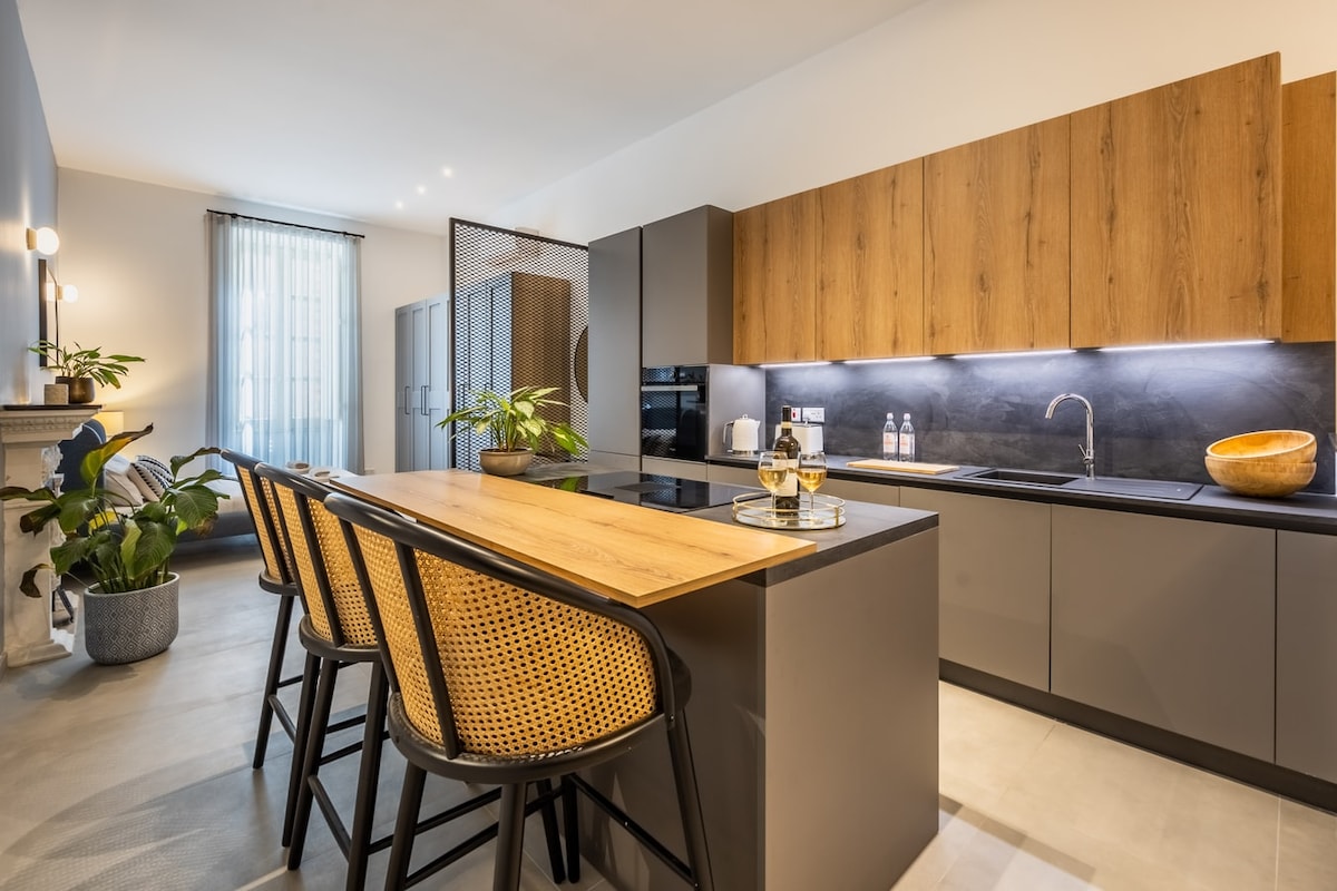 A modern kitchen area features a spacious island with four high-backed chairs. Cabinetry in warm wood contrasts with sleek gray surfaces. Natural light filters in through a large window, illuminating the minimalist design and showcasing a variety of kitchen appliances.