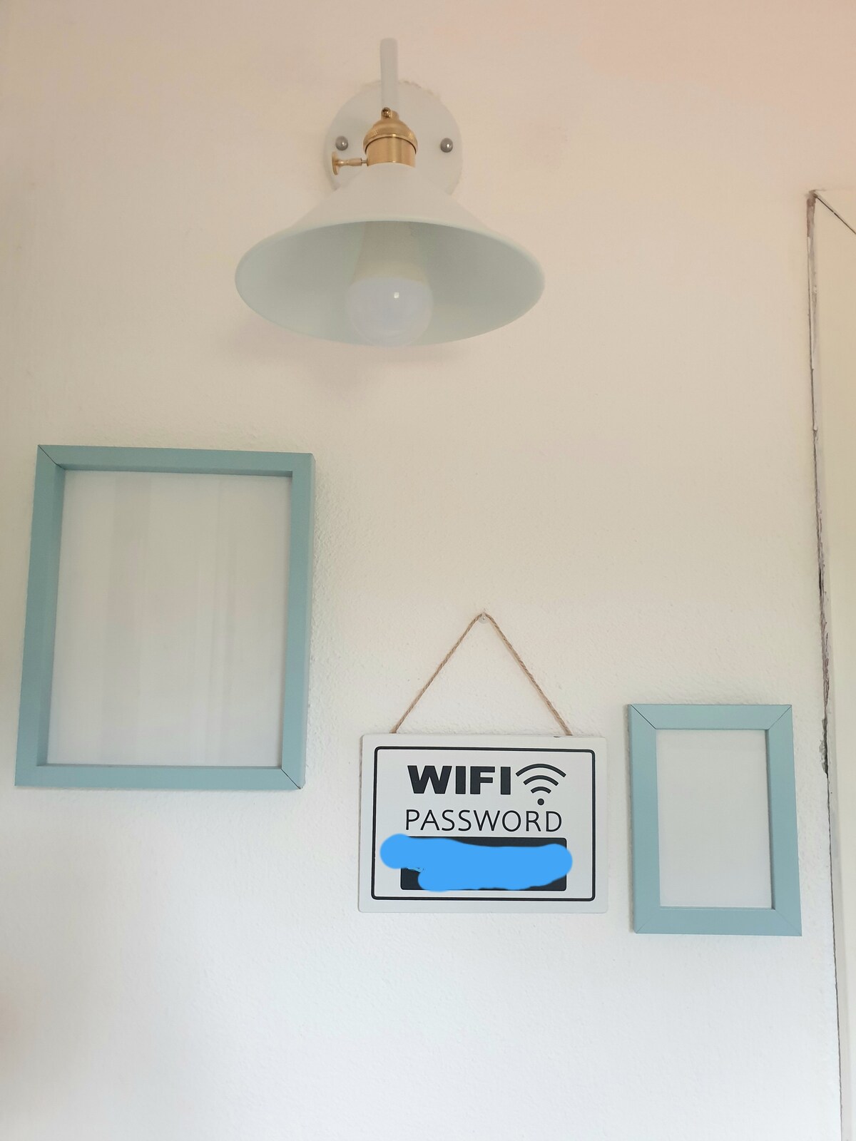 A light fixture is mounted on a wall, providing illumination. Below, a sign displaying the WiFi password is hung on a string. Two empty frames in a pale blue color are positioned nearby, offering a subtle decorative touch.