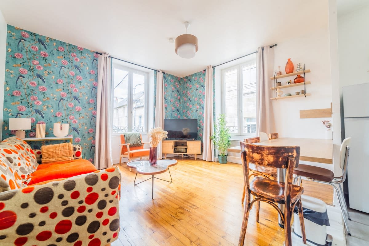 A lively living space features floral-patterned wallpaper and a comfortable couch adorned with colorful polka dots. An entertainment center with a television is positioned against the wall. Large windows provide abundant natural light, complemented by light wood flooring and a dining table set for four.