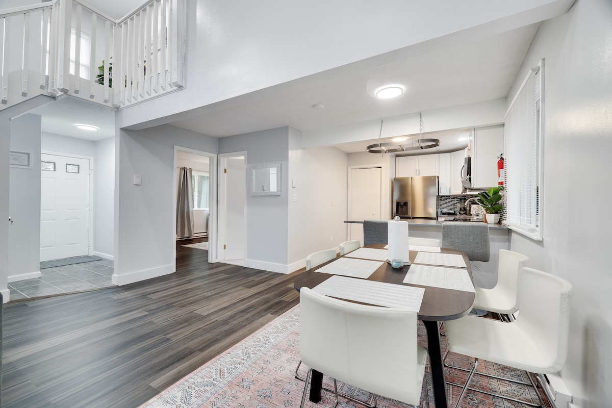 An open living space is displayed, featuring a dining area with a table surrounded by four white chairs. A modern kitchen is visible in the background, equipped with stainless steel appliances. Natural light fills the area through nearby windows, illuminating the light hardwood floor.