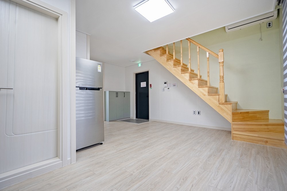 The interior space is characterized by light-colored flooring and a minimalist design. A staircase with wooden banisters leads to the upper level. A silver refrigerator stands against the wall, while an adjacent door provides access to various amenities.