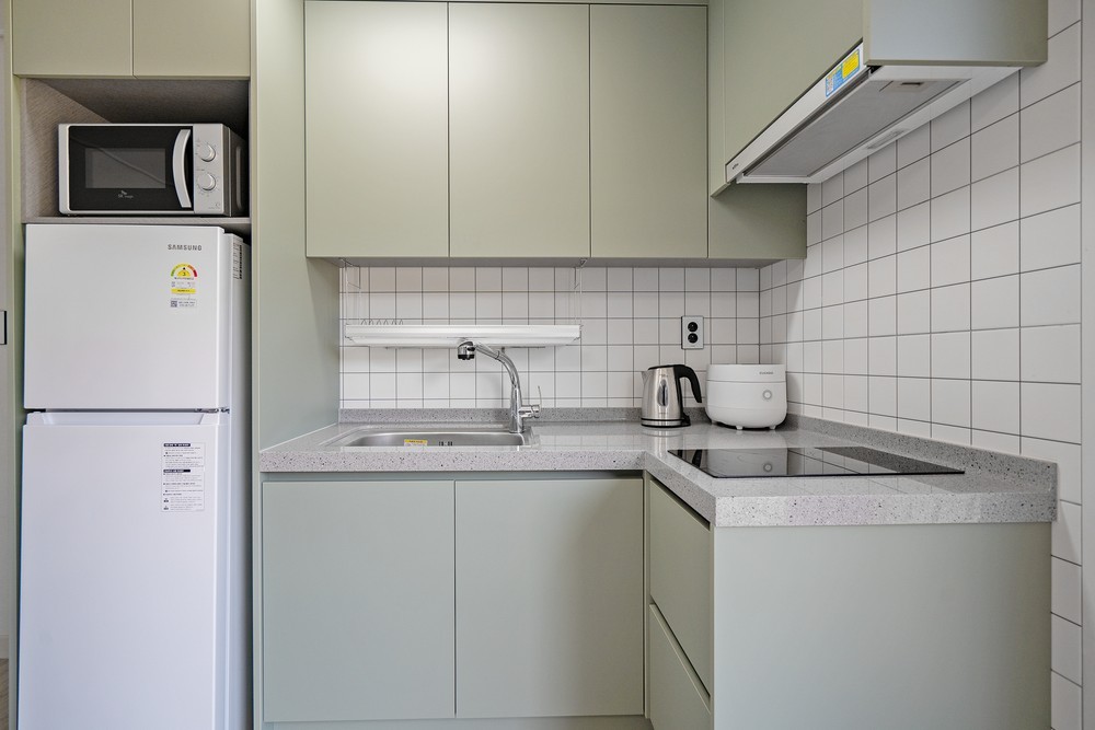 A modern kitchen area features sleek cabinetry in muted green tones, complemented by a light grey countertop. Essential appliances, including a refrigerator, microwave, kettle, and induction stove, are neatly arranged, while white tiles provide a clean backdrop for the functional space.
