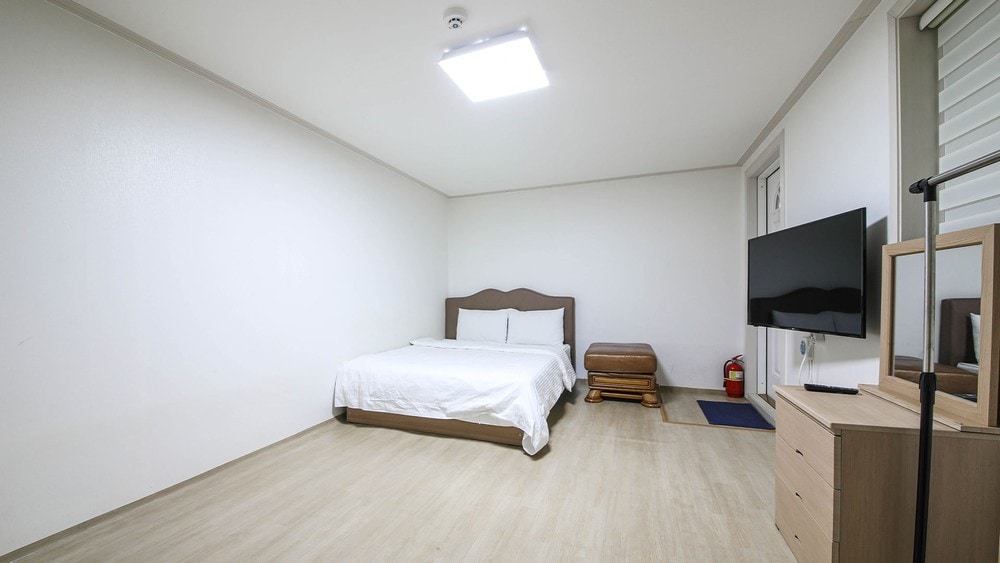 A spacious room features a large double bed against a minimalist backdrop. A television is mounted on the wall opposite the bed. Natural light is provided by a skylight, and a small wooden table is positioned beside the bed. A dresser is visible in the corner.