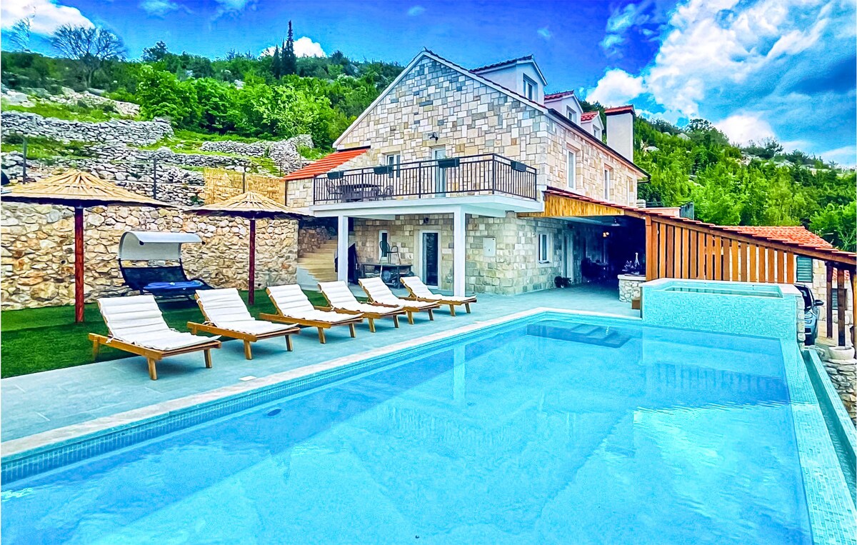 A spacious stone holiday home is surrounded by lush greenery, featuring a private swimming pool. Six loungers are positioned beside the clear water, inviting relaxation. A shaded area provides shelter for seating. The balcony above offers additional outdoor space with views of the landscape.