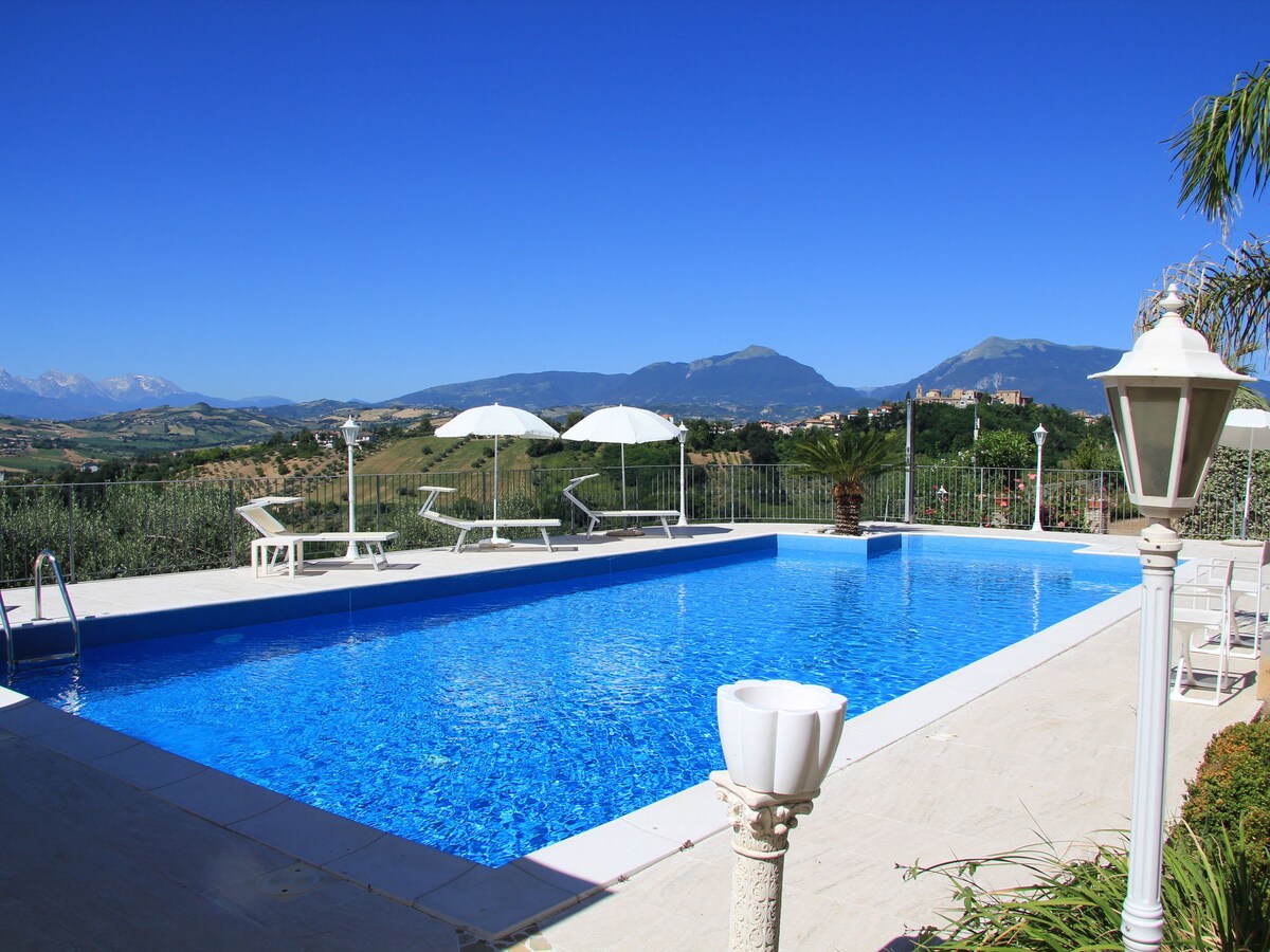 A large swimming pool is set against a backdrop of mountains and clear blue skies. Multiple lounge chairs are arranged around the pool area, accompanied by white umbrellas for shade. Lush greenery can be seen in the surrounding landscape.