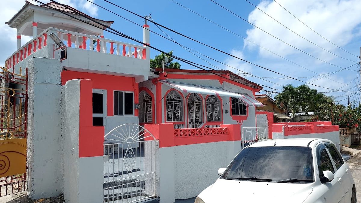 The exterior of the house features a combination of light and vibrant coral paint, with decorative wrought iron accents. A gated entrance leads to a welcoming porch area. The vehicle parked in front indicates convenient accessibility.