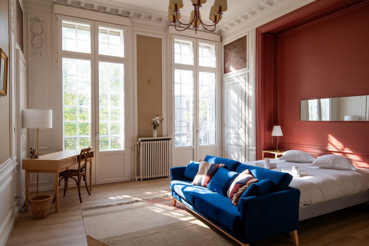 A spacious living area is illuminated by natural light streaming through large windows, reflecting off the elegant furnishings. A blue sofa provides comfortable seating, complemented by a small desk and chair. The double bed is adorned with simple white linens, and a decorative mirror enhances the room's dimension.
