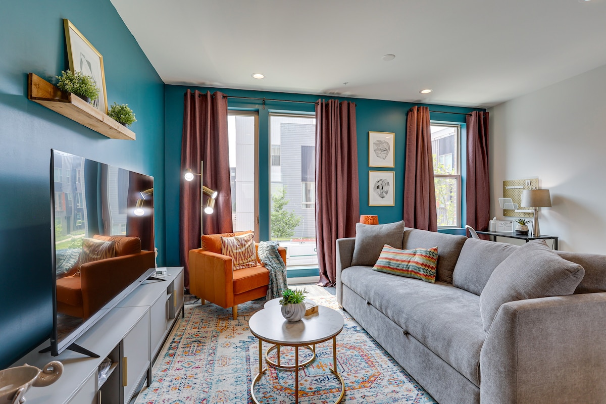 A cozy living area features a spacious gray sofa and an orange accent chair. Soft light from the large glass doors illuminates the room, where a round coffee table sits atop a decorative rug. Dark curtains frame the windows, enhancing the inviting atmosphere.