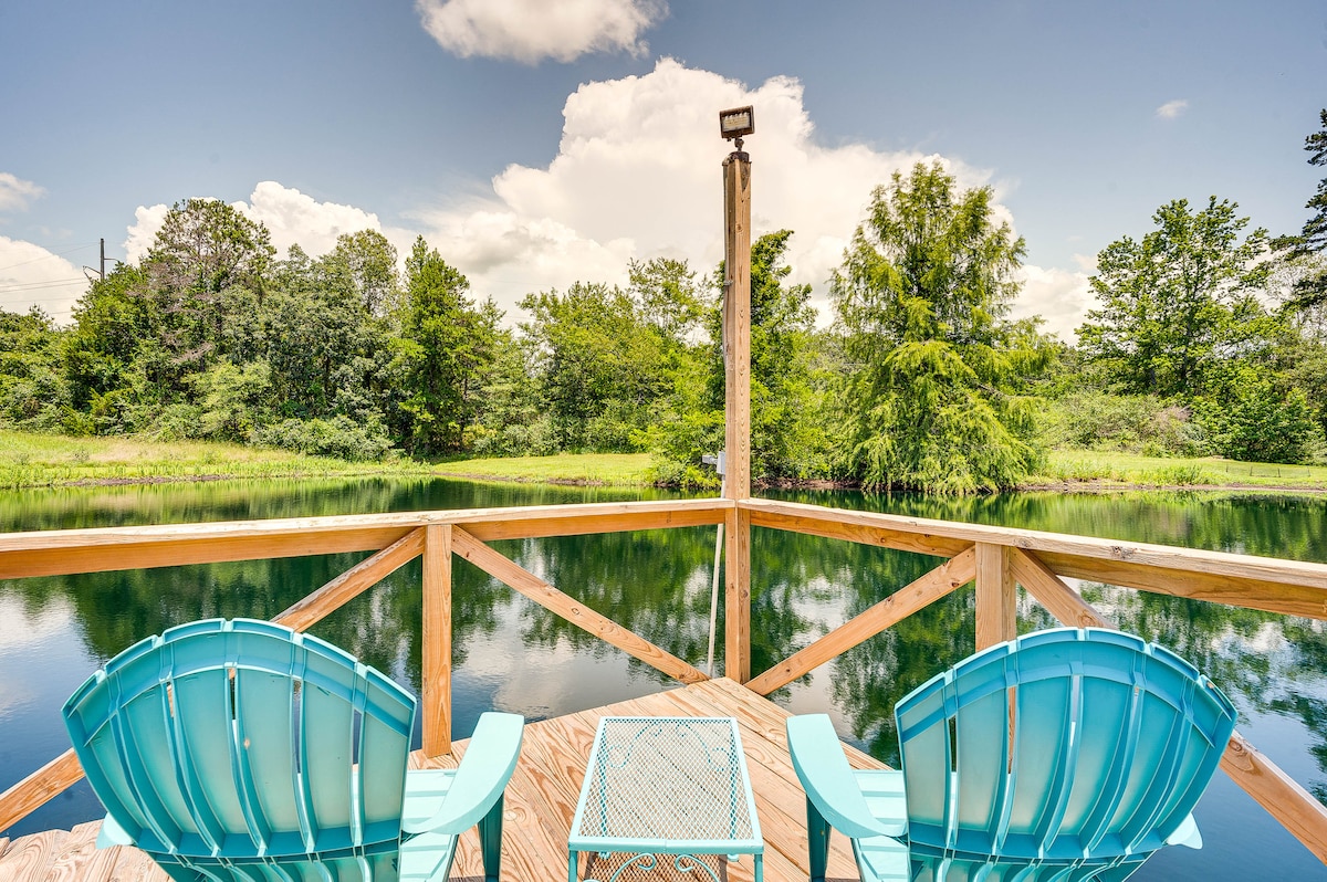 A private fishing pier extends over a serene pond, flanked by lush green trees and clear skies. Two turquoise chairs are positioned beside a small table, offering a peaceful spot to enjoy nature's beauty. Reflections of the surrounding greenery are visible on the water's surface.