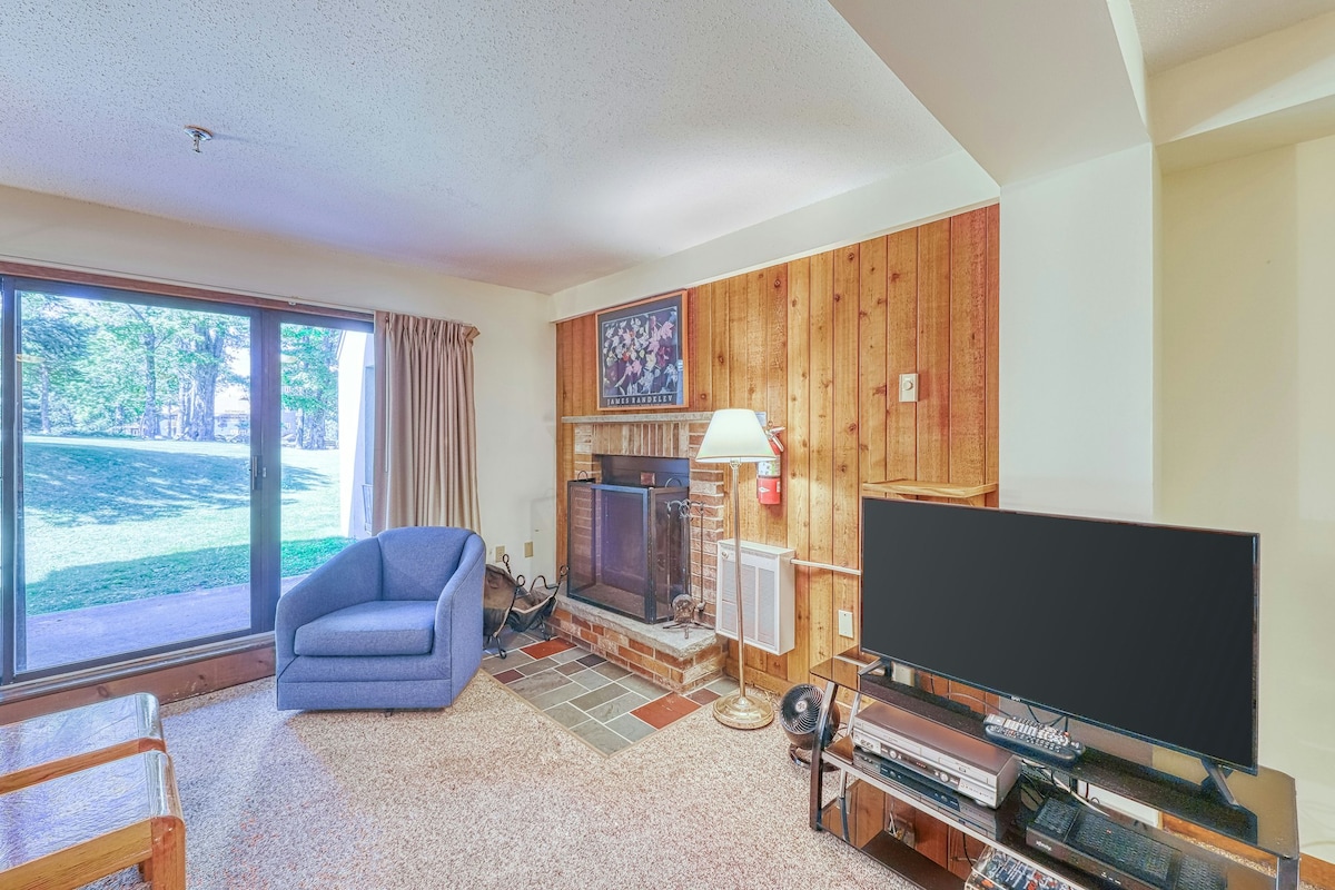 A comfortable living space features a large window overlooking a private yard, allowing natural light to brighten the room. A blue chair is positioned near the stone fireplace, while a television and entertainment center occupy one corner, providing a cozy area for relaxation.