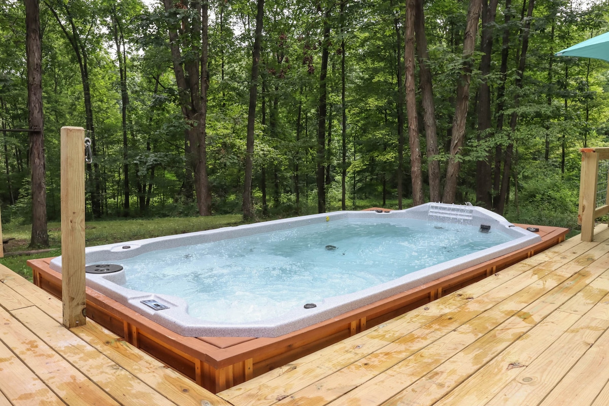An outdoor heated swim spa is set on a wooden deck, surrounded by lush greenery. The water is gently bubbling, inviting relaxation in a serene forested setting.
