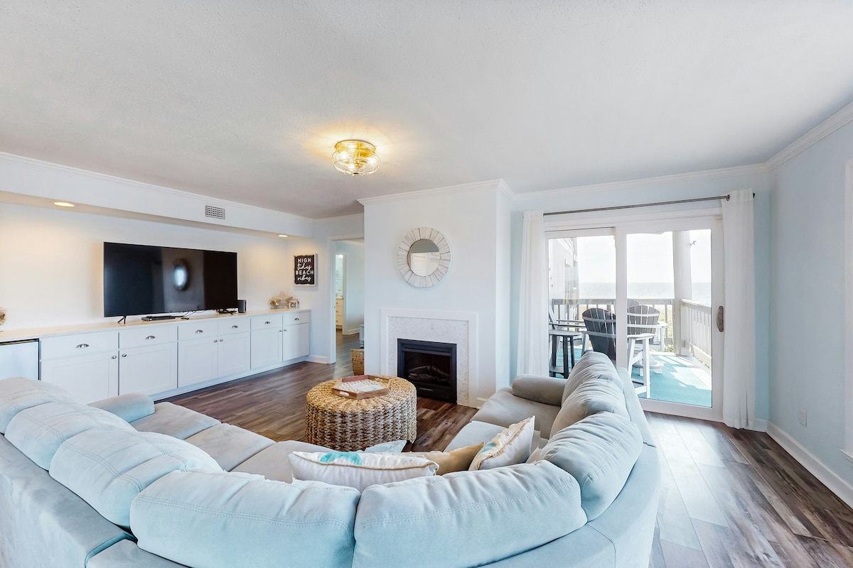 A spacious living area features a plush sectional sofa, centered around a round coffee table made of natural materials. A large TV is mounted on a wall unit. Light pours in through sliding glass doors, leading to a private balcony with ocean views, while a fireplace adds warmth.