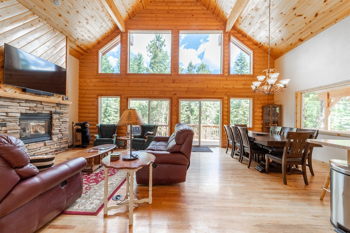 A spacious living area features vaulted ceilings with large windows offering views of surrounding trees. Leather seating arrangements are positioned near a stone fireplace. A wooden dining table accommodates multiple guests, while natural light enhances the warm tones of the wood walls and floor.