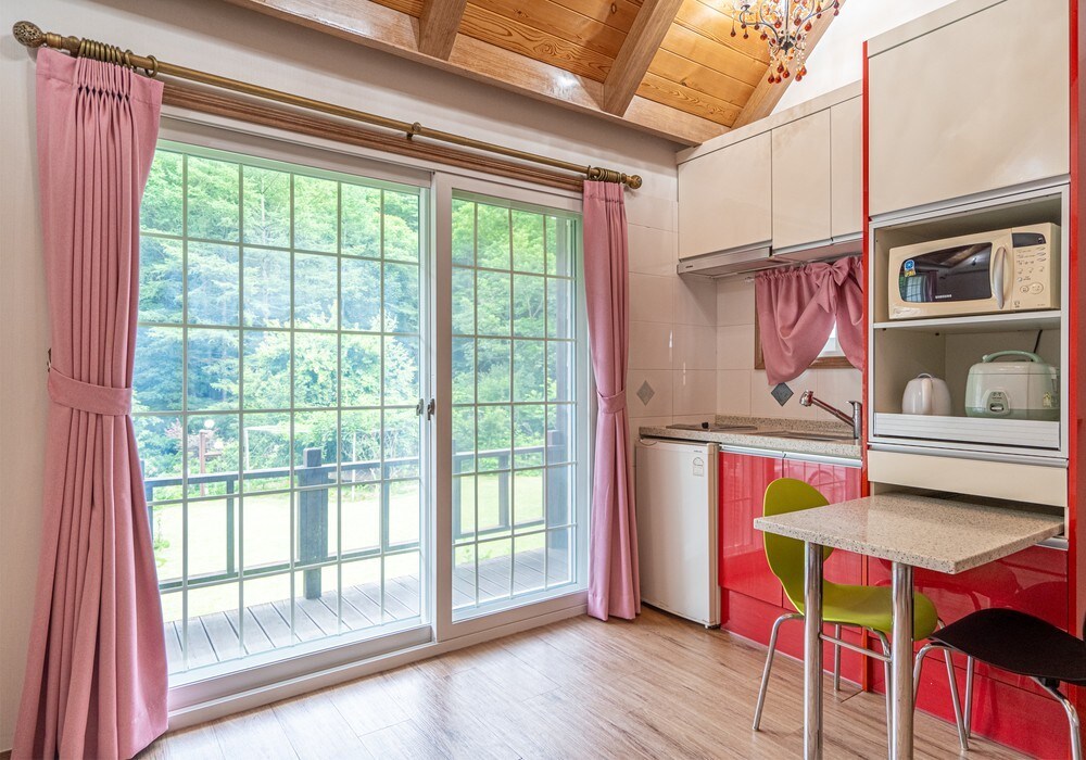 The kitchen area features modern cabinetry and a small dining table with two chairs. Natural light floods in through large glass doors, revealing a view of a lush green landscape. Soft pink curtains frame the windows, adding a touch of warmth to the space.