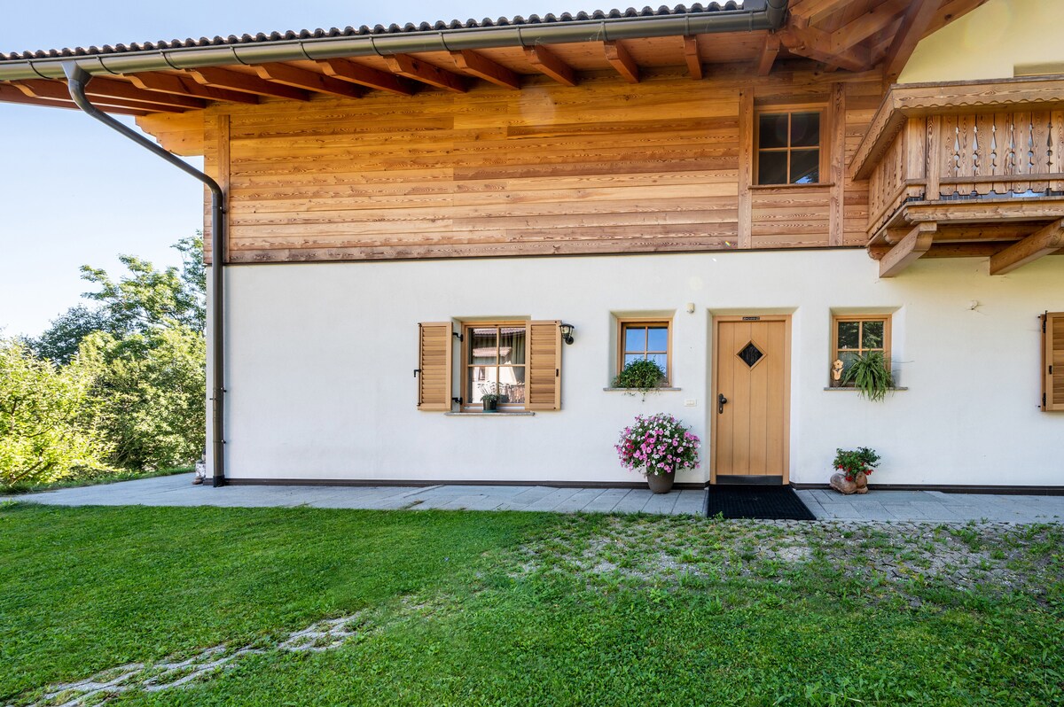 A wooden exterior features a blend of natural textures and colors. The entrance, framed by flower pots, is complemented by a welcoming door. Windows with shutters allow for natural light, while a grassy area surrounds the building, creating a serene outdoor environment.