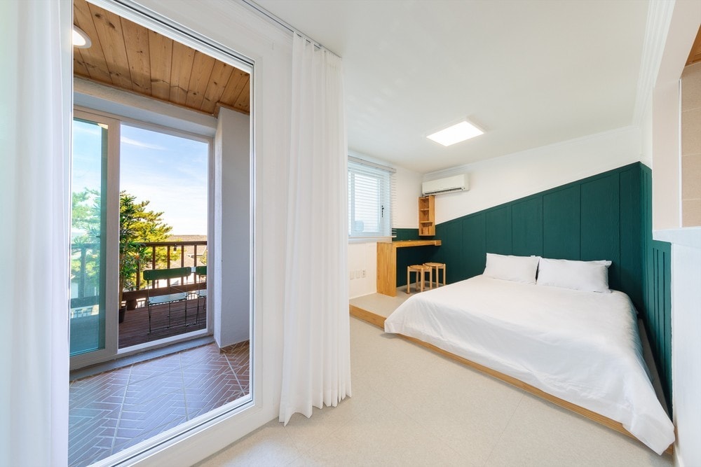 A well-lit room features a spacious bed adorned with crisp white linens. A small desk and two chairs are positioned against a wall, and a sliding glass door opens to a private balcony, allowing natural light to flow in from the outdoor space.