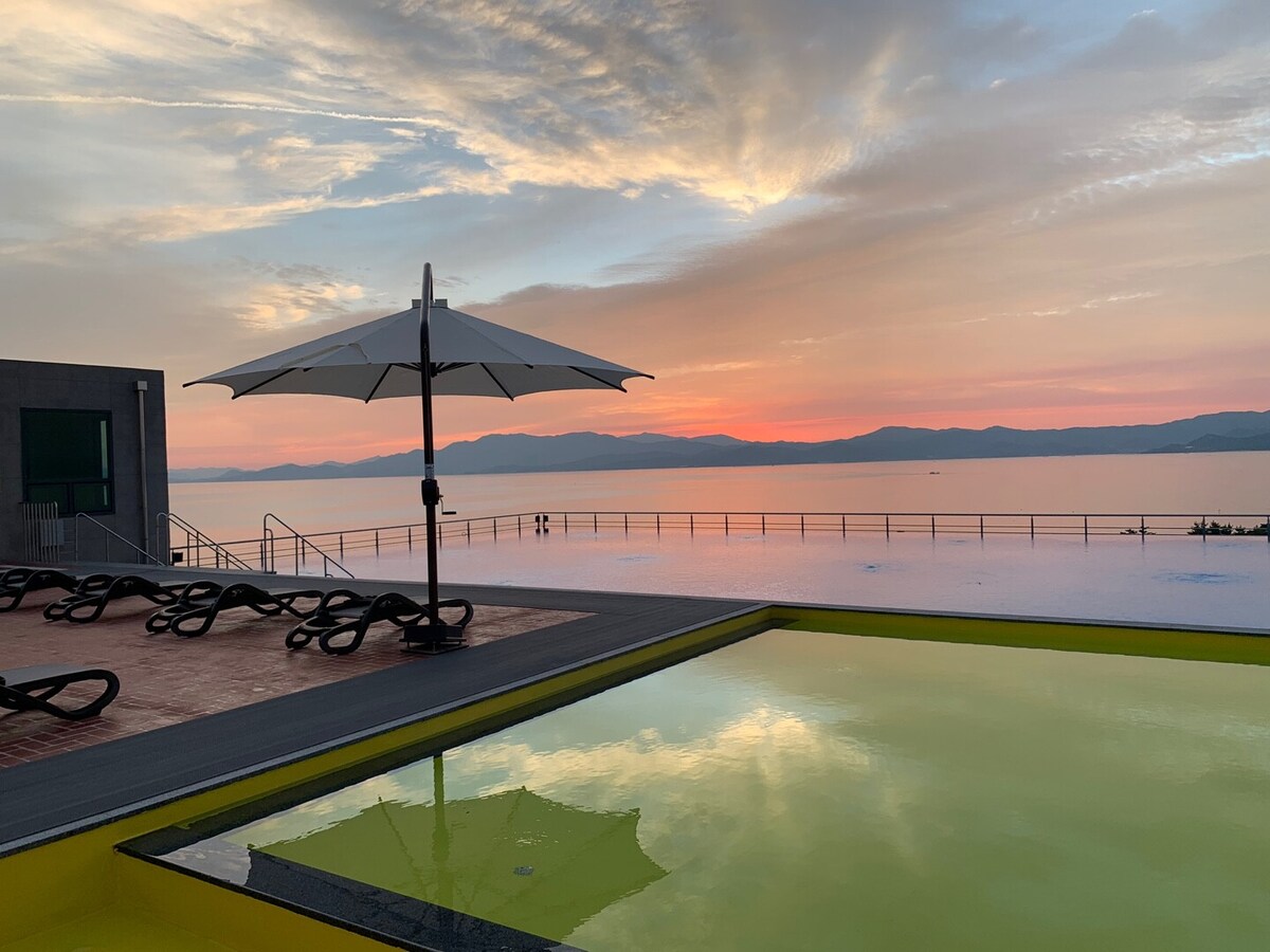 An infinity pool is visible, reflecting soft hues of a sunset against a serene backdrop of mountains and water. A large umbrella provides shade over a sun lounger, complementing the tranquil setting. The scene evokes a sense of calm and relaxation.