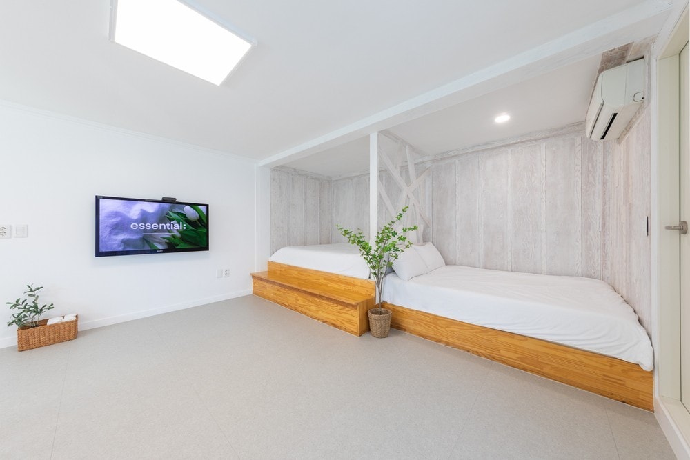 A bright sleeping area features two elevated beds made of light wood, providing a minimalist aesthetic. A television mounted on the wall is positioned opposite the beds, while a planter with greenery adds a touch of nature to the space.