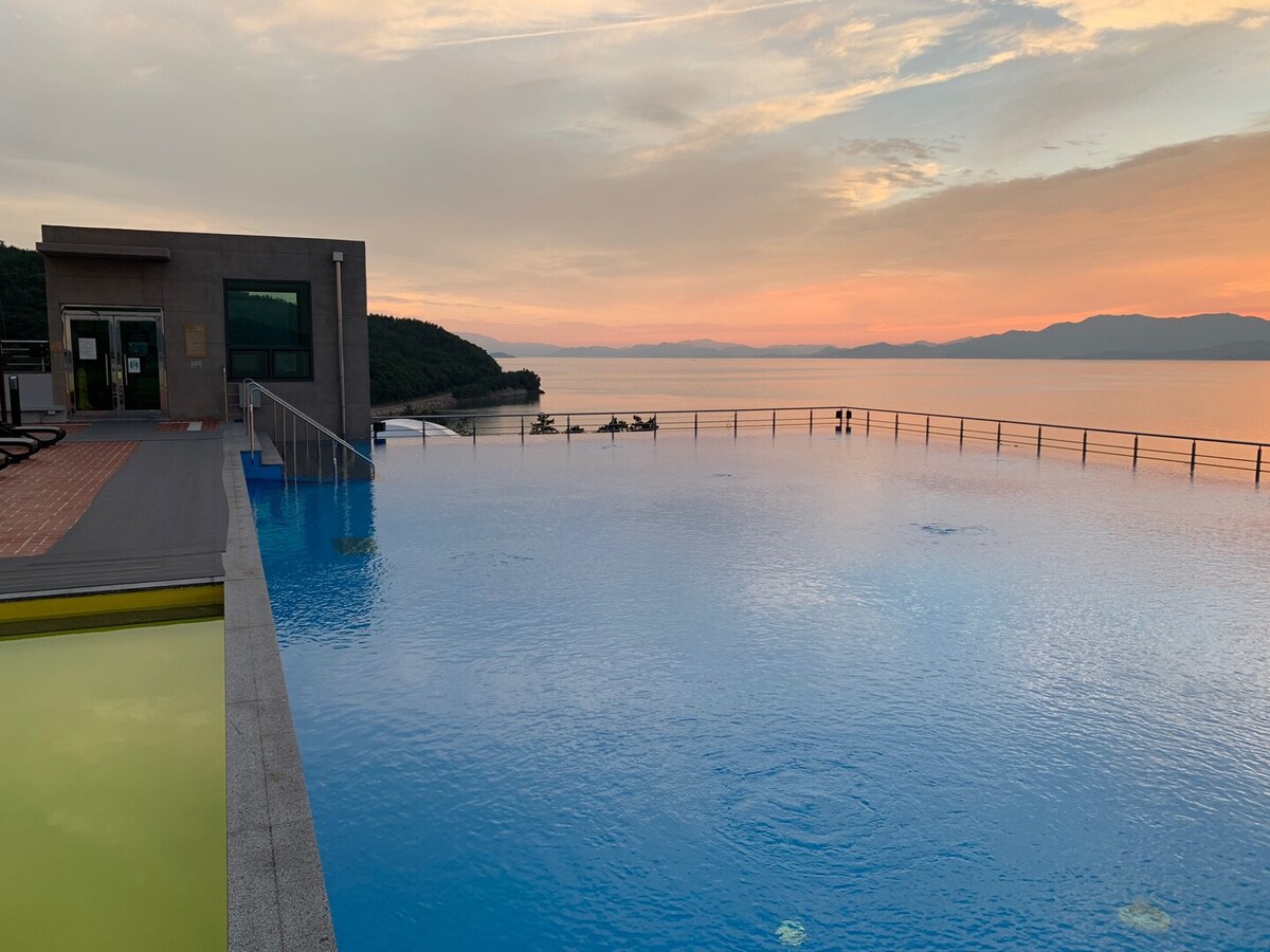 An infinity pool stretches towards the horizon, reflecting the soft hues of the sunset above. The serene water appears calm, with a distant view of the mountains and coastline. Comfortable seating is visible nearby, creating a relaxing atmosphere for guests.