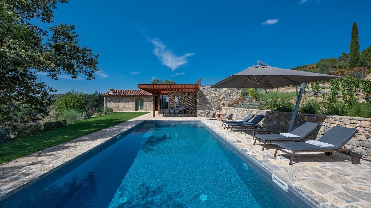 A private pool is surrounded by stone paving and a well-maintained lawn. Sun loungers with cushions are positioned near a large parasol. A shaded seating area with a roof structure is visible in the background, blending with the lush greenery and Tuscan landscape.