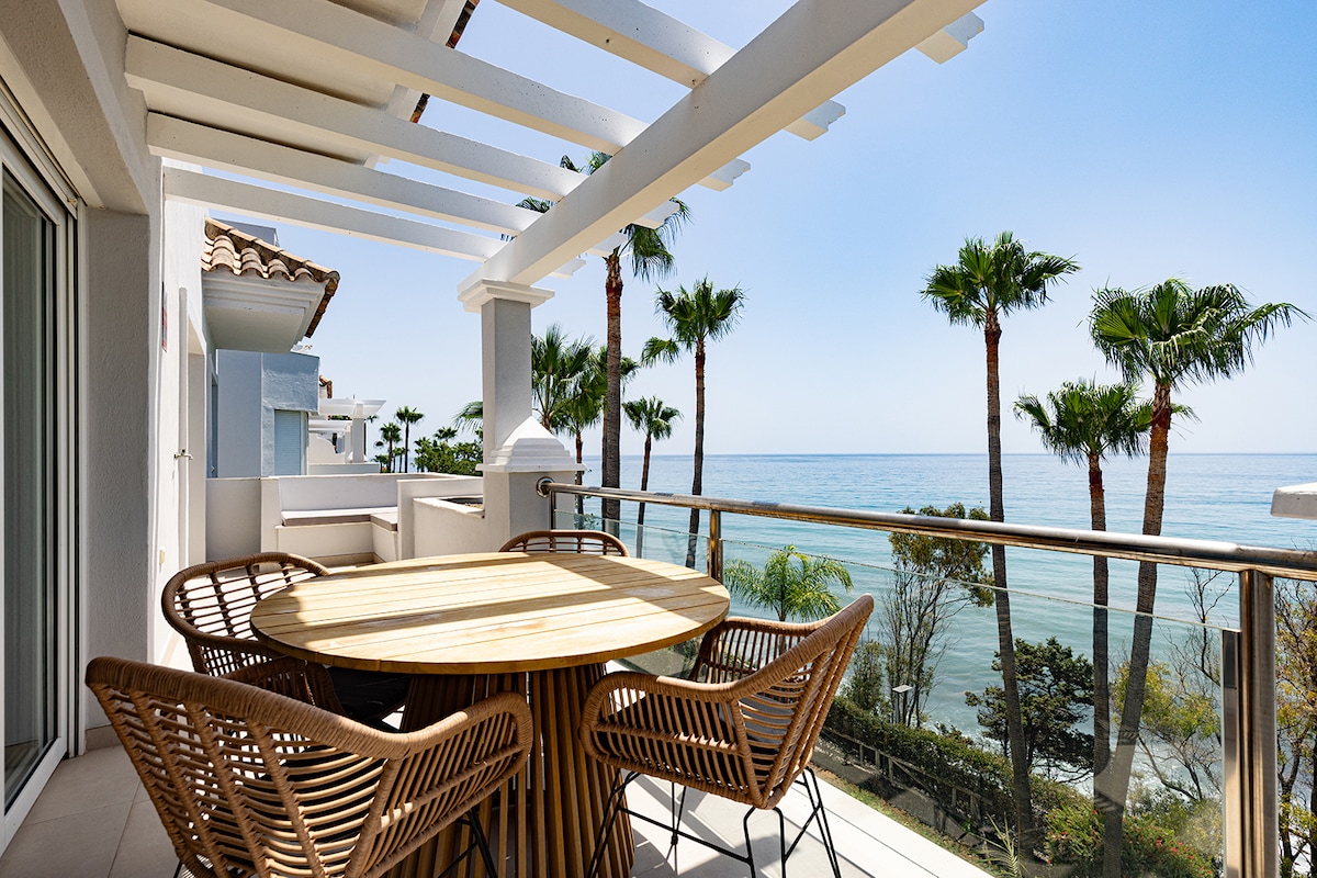 A spacious outdoor terrace is equipped with a round wooden table surrounded by stylish woven chairs. A scenic view of palm trees and the calm sea is visible in the background, creating a tranquil setting for outdoor relaxation.