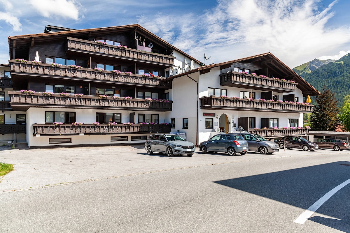 The building features a classic alpine architecture with multiple balconies adorned with flower boxes. A spacious parking area is located in the front, accommodating several vehicles, while lush greenery and mountains can be seen in the background under a partly cloudy sky.