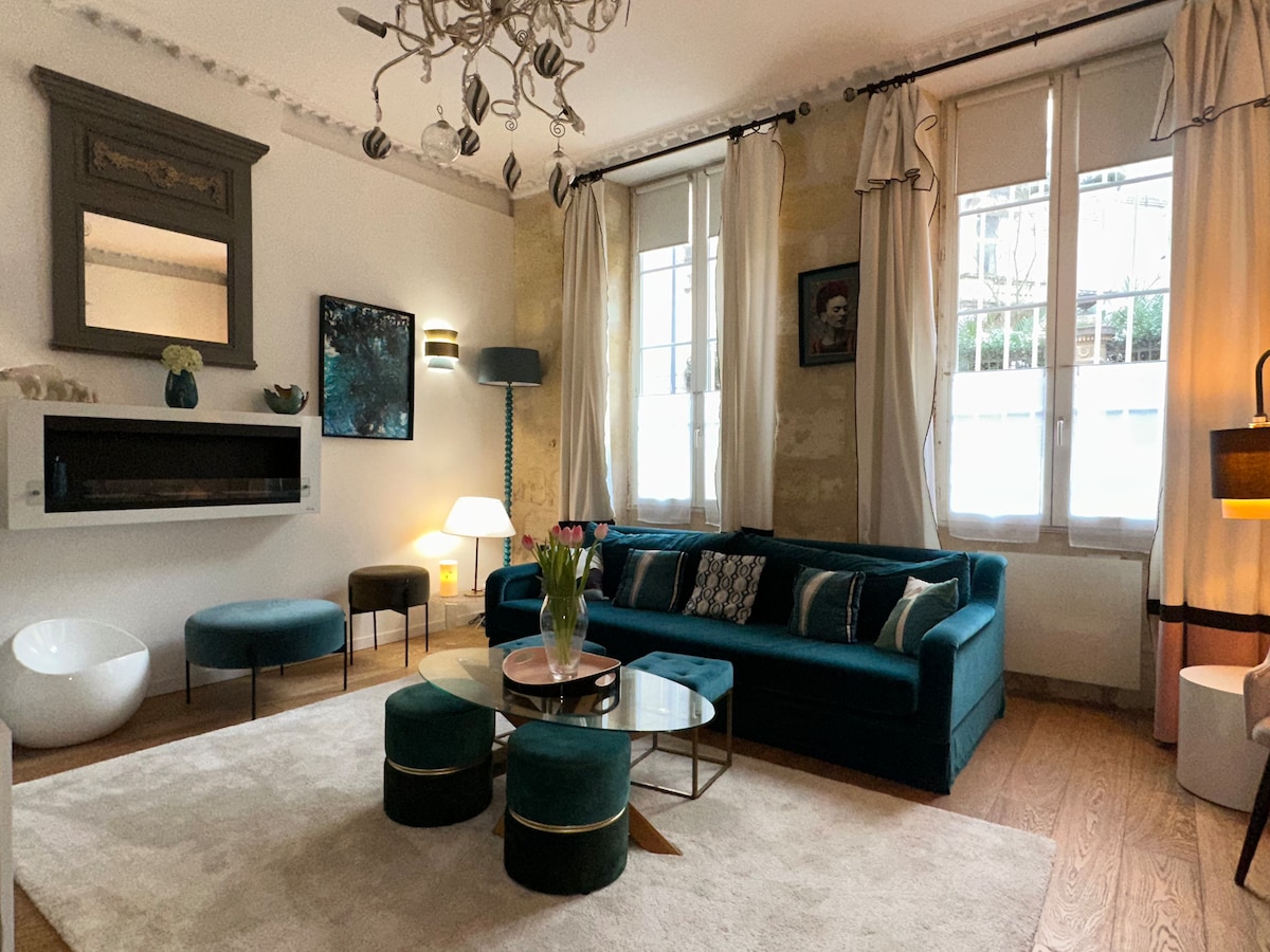 The living area features a turquoise sofa surrounded by round ottomans and a glass coffee table. Natural light enters through large windows, while decorative elements include a modern chandelier and framed artwork. A calming atmosphere is created with neutral tones and soft textures.
