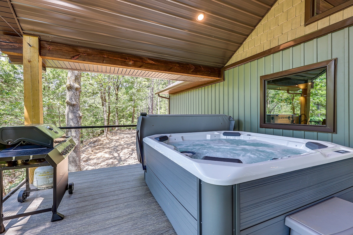 A private hot tub is positioned on a covered deck, surrounded by lush greenery. Nearby, a gas grill is available for outdoor cooking. Natural light filters through the wooden beams above, creating a peaceful outdoor setting.
