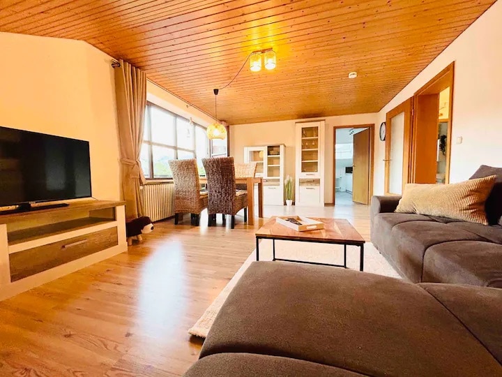 A spacious living area features a large sectional sofa and a mounted television. Natural light enters through the large windows, complemented by warm wooden paneling on the ceiling. A dining table with four chairs can be seen in the background, along with an inviting open layout.