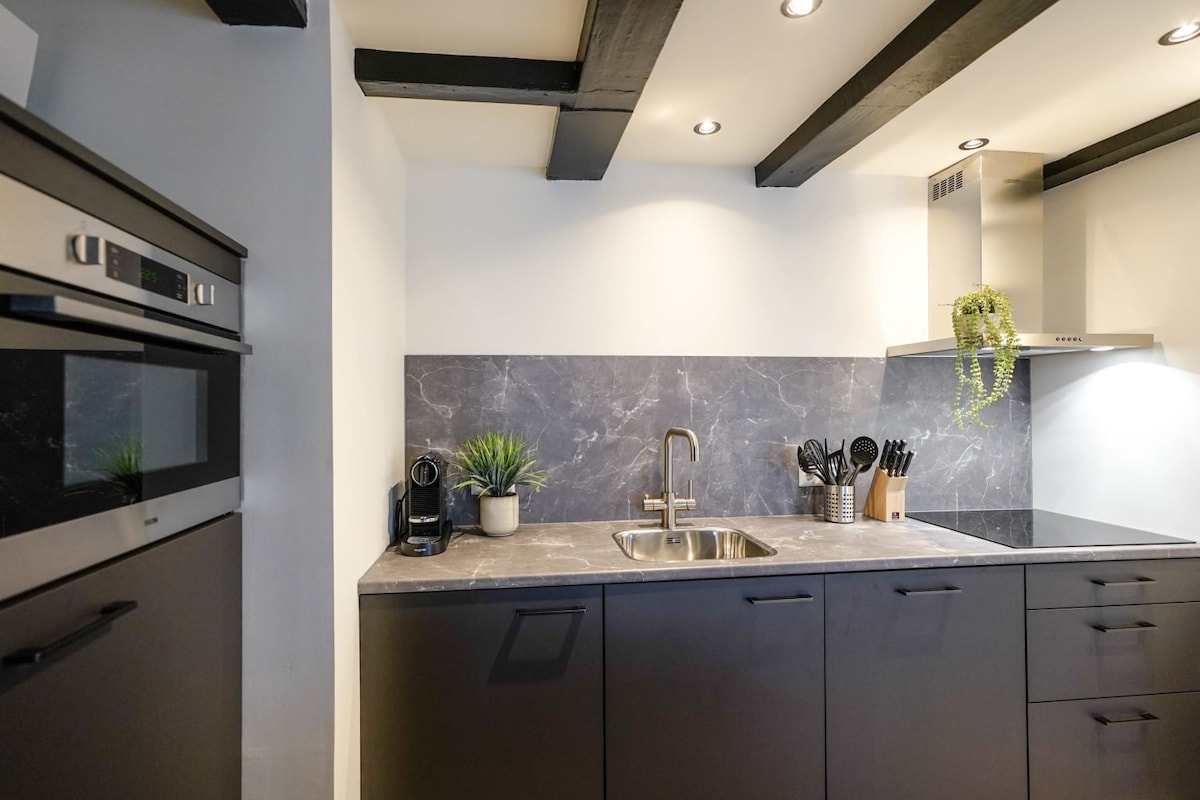 A modern kitchen showcases sleek black cabinetry, accented by a marble countertop. Essential appliances including an oven, coffee maker, and various utensils are neatly arranged. Soft lighting highlights the elegant design, while a small plant adds a touch of greenery to the space.
