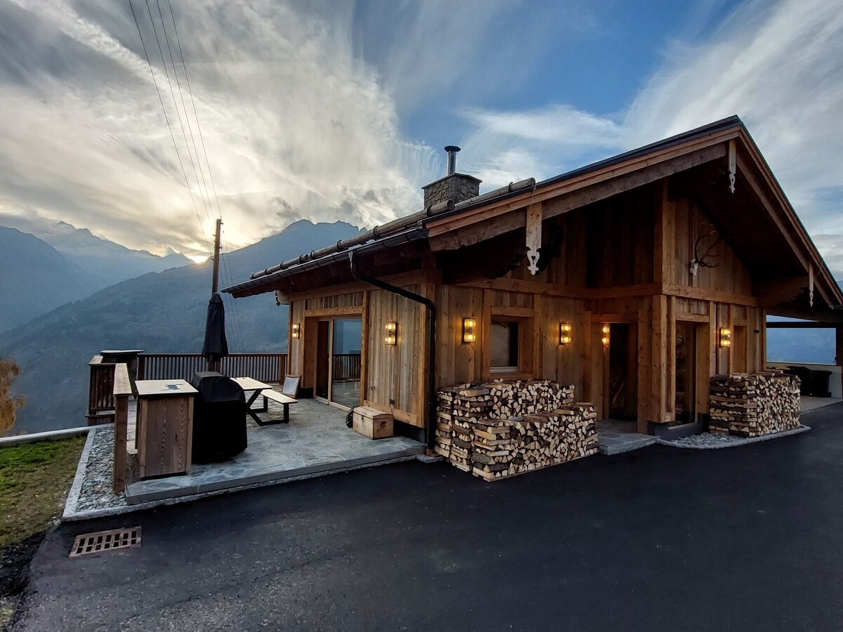 A rustic chalet exterior is presented, featuring wooden construction and large windows that showcase the mountain views. Stacked firewood is neatly arranged along the side. An outdoor seating area is visible, with a sun umbrella nearby, against a backdrop of mountains and a cloudy sky.