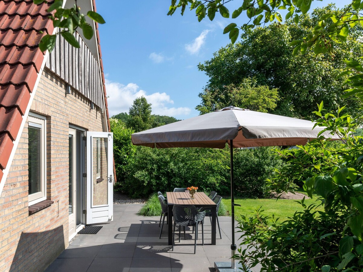 An outdoor terrace is shown featuring a shaded table with four chairs. A parasol provides shelter from the sun, and lush greenery surrounds the area, enhancing the sense of privacy. The house's exterior displays a combination of brick and wooden features under a sloped roof.