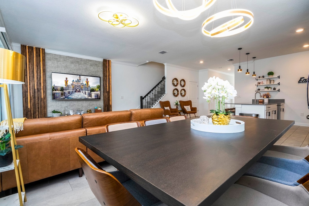 A spacious dining area features a dark wooden table surrounded by contemporary chairs. A large, inviting sofa is visible in the background, along with a decorative wall display and a mounted television. Soft lighting from modern fixtures creates a warm ambiance.
