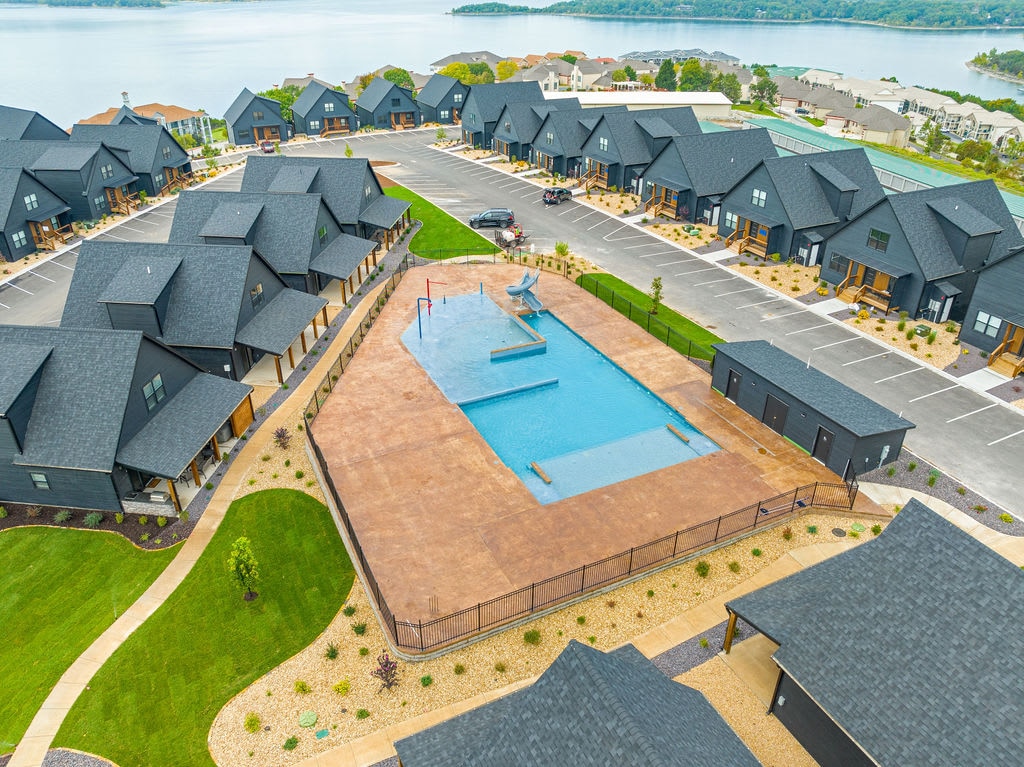 An aerial view captures the pool area surrounded by neatly arranged lodge-style buildings. The clear water of the pool contrasts with the textured surface surrounding it. Vast open spaces and walking paths integrate green landscaping, leading toward scenic views of the lake in the background.