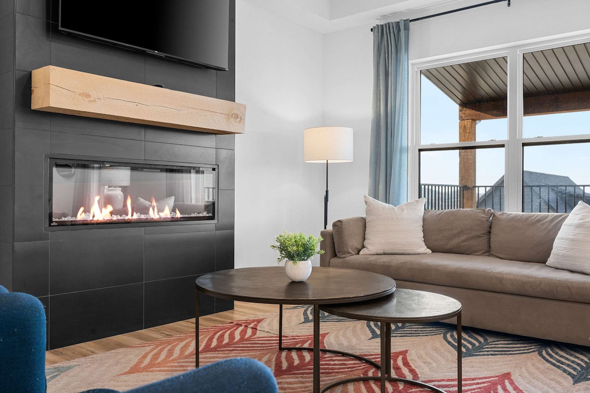 A modern living area features a sleek electric fireplace set within a black wall. A light-colored couch is positioned nearby, complemented by accent chairs and a round coffee table. Large windows provide views of the outdoor deck and bring in natural light, while soft curtains frame the space.