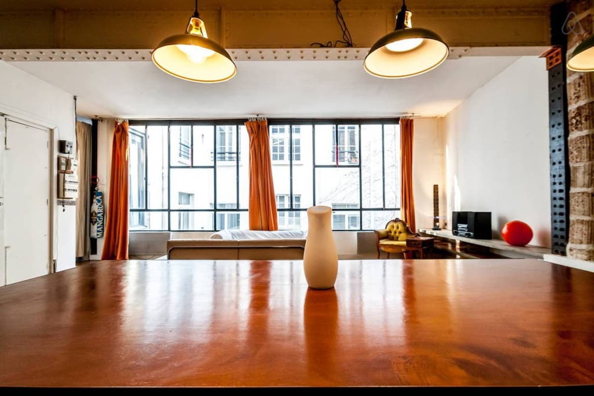 The image showcases a spacious loft with large, well-lit windows framed by orange curtains. A centrally positioned wooden table is highlighted, with a simple white vase resting atop. In the background, a cozy bed and a desk with decorative items can be seen, contributing to the room's modern character.