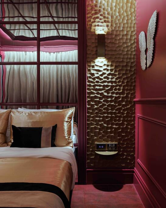 A stylish sleeping area features a queen-sized bed with luxurious bedding in cream and gold tones. Textured wall panels emit soft light, while a decorative wing sculpture adds a unique touch to the vibrant red walls.