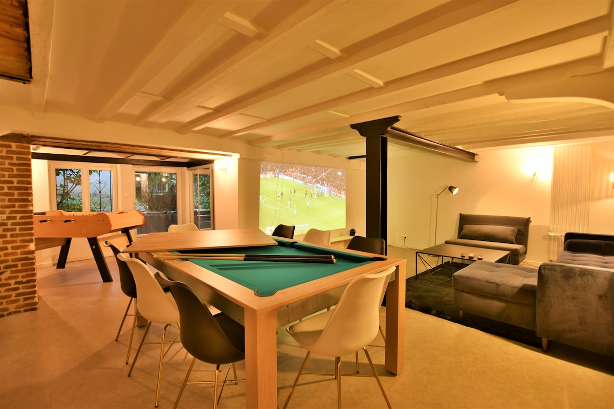 A recreation area features a billiard table and a foosball table against a rustic brick wall. A large screen is mounted for movie viewing, with a comfortable sofa positioned for optimal viewing. Bright lighting enhances the spacious feel of the room.