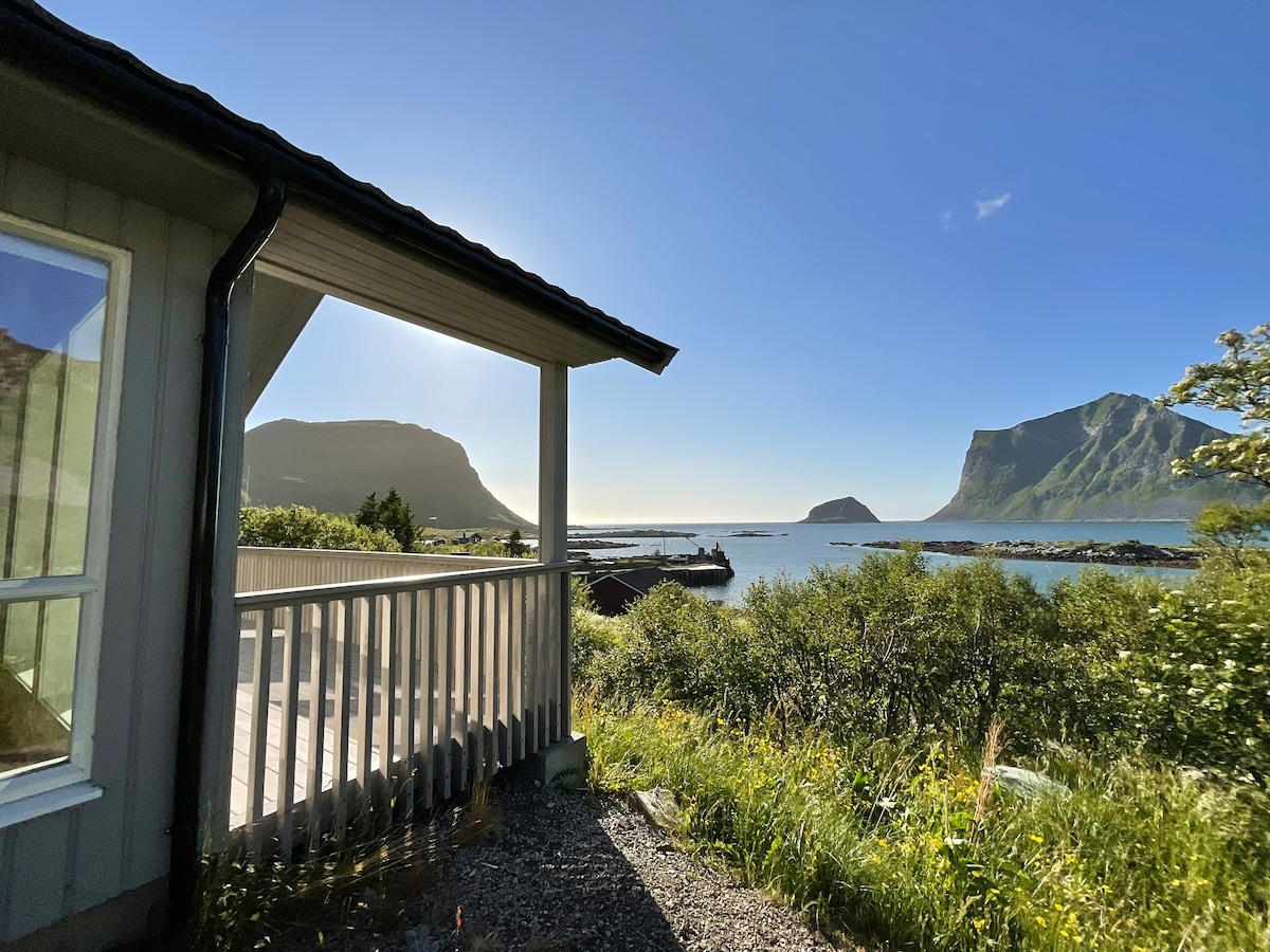 A view from the cabin's terrace captures the surrounding mountains and the sea. The wooden railing frames the landscape, complemented by lush greenery in the foreground. Bright sunlight enhances the natural beauty, creating a serene atmosphere along the coastline.