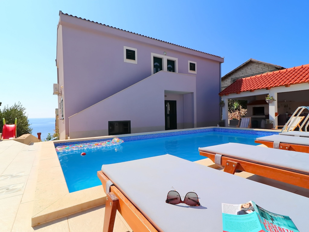 The outdoor space features a private heated swimming pool surrounded by lounge chairs. A bright, modern building is visible in the background, accompanied by clear blue skies and glimpses of the sea. A colorful inflatable float rests in the pool, enhancing the inviting atmosphere.