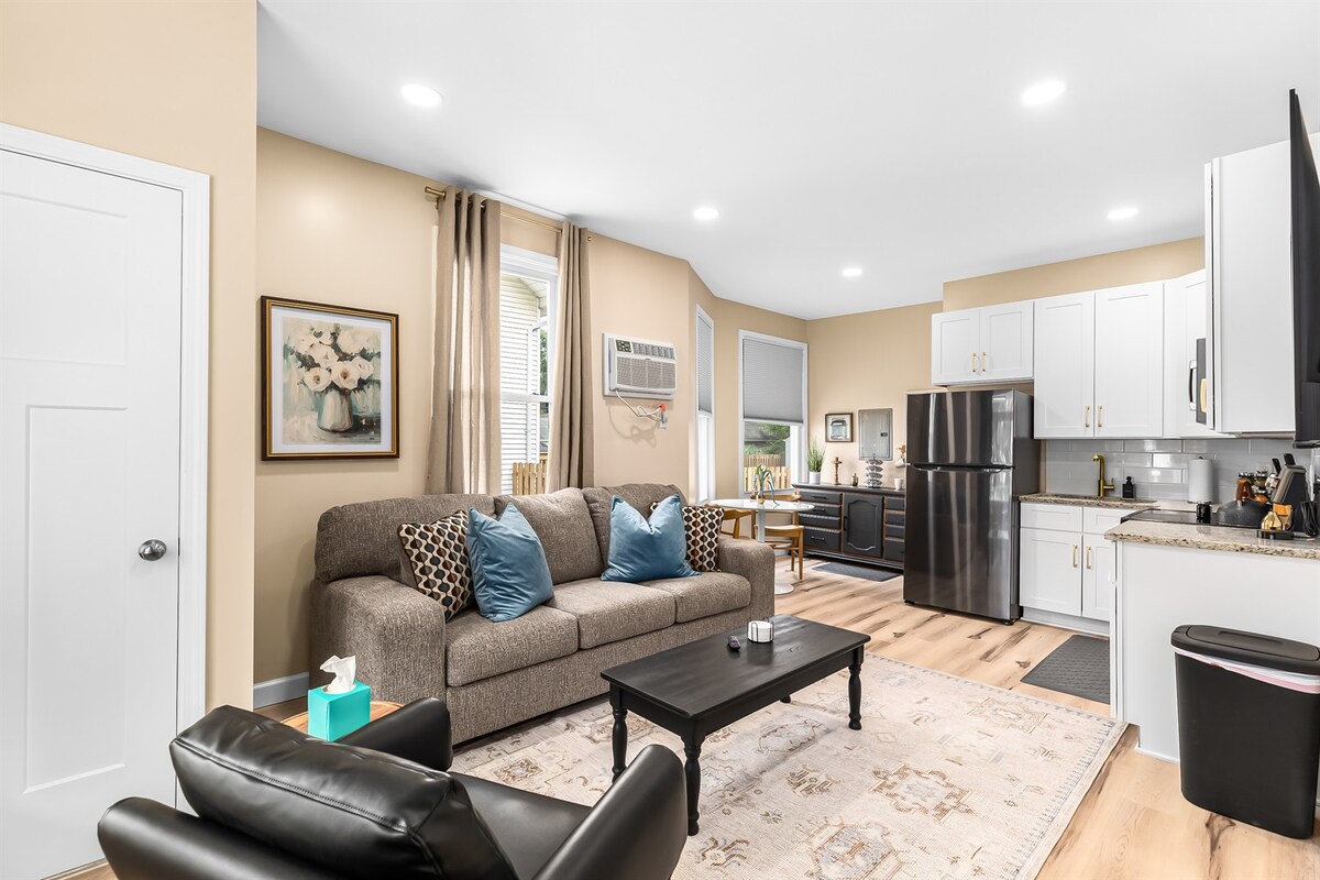A cozy living area features a comfortable sofa adorned with decorative pillows and blankets. A dark coffee table sits in front of the sofa, with a flat-screen TV mounted on the wall. The open kitchen is equipped with modern appliances and white cabinetry, complemented by warm lighting.