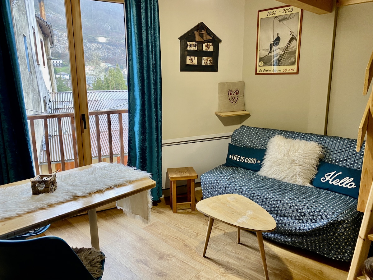 A cozy living area is presented, featuring a comfortable sofa with a decorative blue cover and pillows. A wooden coffee table occupies the center, complemented by a small side table. Natural light streams through glass doors, leading to a scenic view outside.