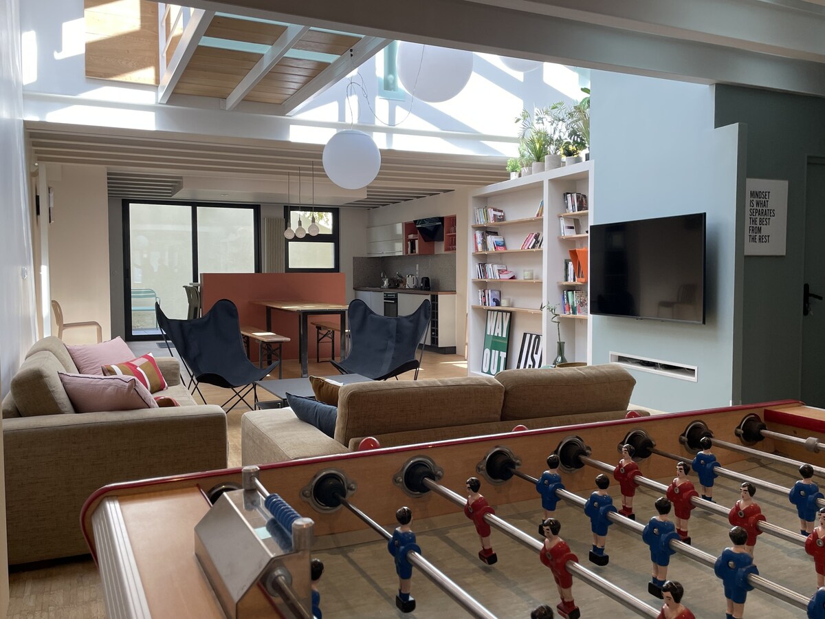 A spacious living area features comfortable sofas and modern armchairs, arranged around a central coffee table. A foosball table is visible in the foreground. The open kitchen and dining space, along with shelves filled with books, are softly illuminated by natural light from large windows.