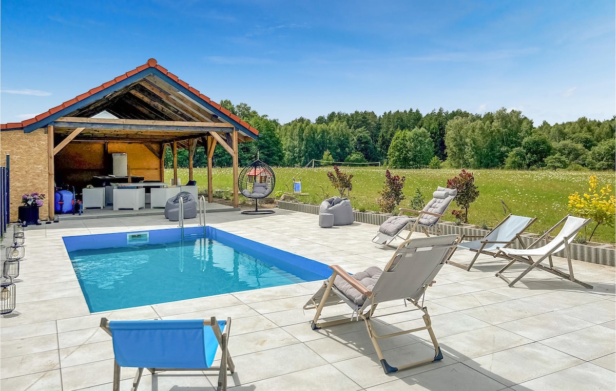 The outdoor area features a refreshing pool surrounded by several lounge chairs and a shaded patio with a structural roof. A grassy field is visible in the background, along with greenery accentuating the peaceful surroundings.