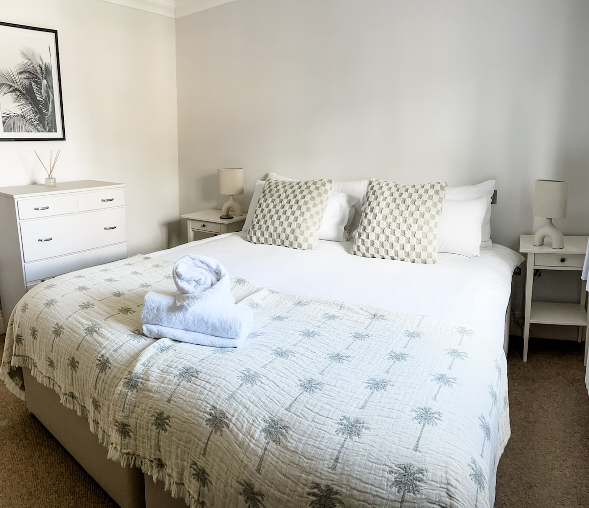 A well-appointed bedroom features a spacious bed with a soft, patterned blanket draped across it. Two bedside tables with lamps flank the bed, while a framed artwork and a minimalist decorative item are displayed on the wall.