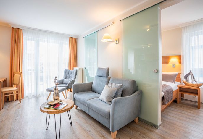 A modern living space is highlighted by a grey sofa and wooden coffee table. Two armchairs are positioned nearby, while a partition with sliding glass panels separates the sleeping area in the background. Natural light streams through sheer curtains, creating a bright and airy atmosphere.