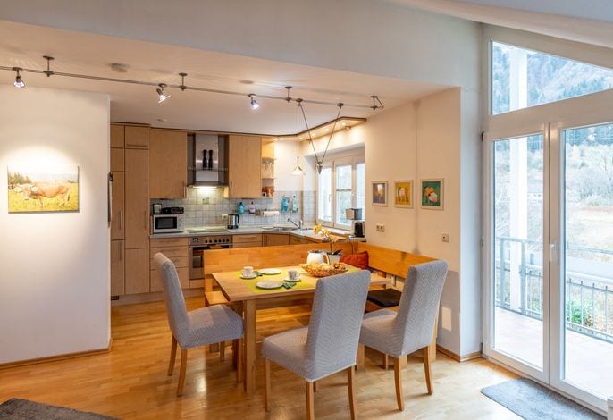 A well-equipped kitchen and dining area is presented, featuring a wooden dining table surrounded by upholstered chairs. Modern appliances, including a microwave and oven, are visible along with vibrant wall art. Large windows provide natural light and access to a balcony with scenic views.
