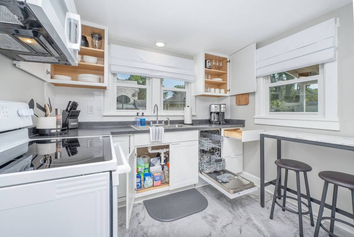 The kitchen features modern cabinetry and ample counter space. Appliances include an oven, microwave, and coffee maker. Open shelves display dishware, while a dishwasher is visible with its door open. A small table with two stools offers additional seating for meals or preparation.